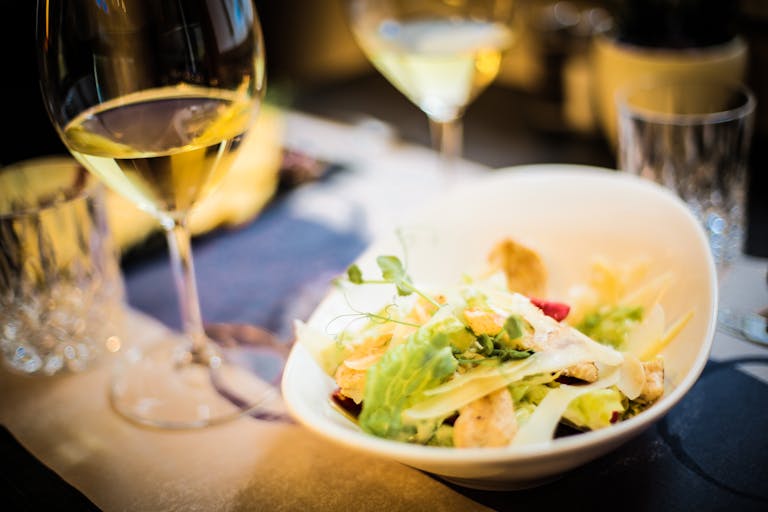 High angle of tasty salad made of fresh vegetables and green herbs placed on table with glass of wine in luxury restaurant